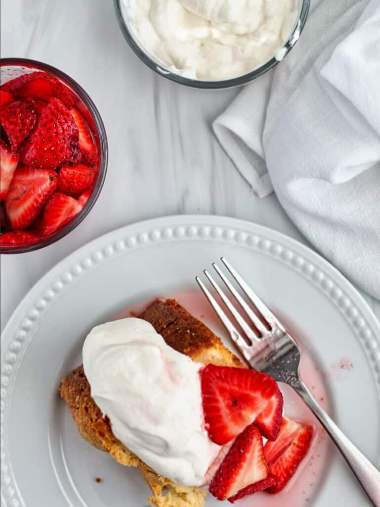 overhead view of pound cake slice on white plate with strawberries and whipped cream with bowls of strawberries and whipped cream and a white towel in the background.