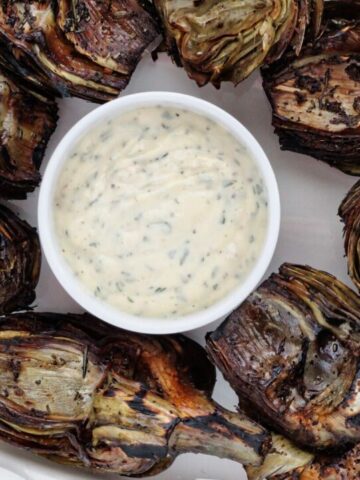 overhead view of fire roasted artichokes on white platter with garlic aioli in the middle