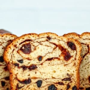 slice of gf cinnamon raisin bread standing upright on white platter with cut open loaves behind it.