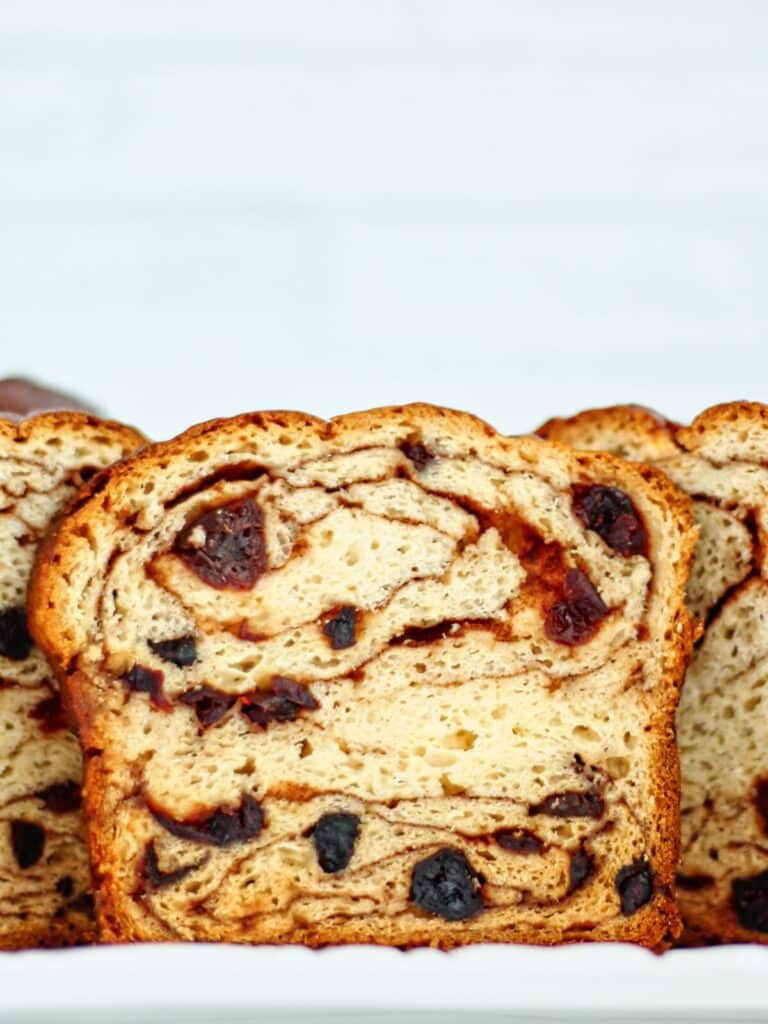 slice of gf cinnamon raisin bread standing upright on white platter with cut open loaves behind it.