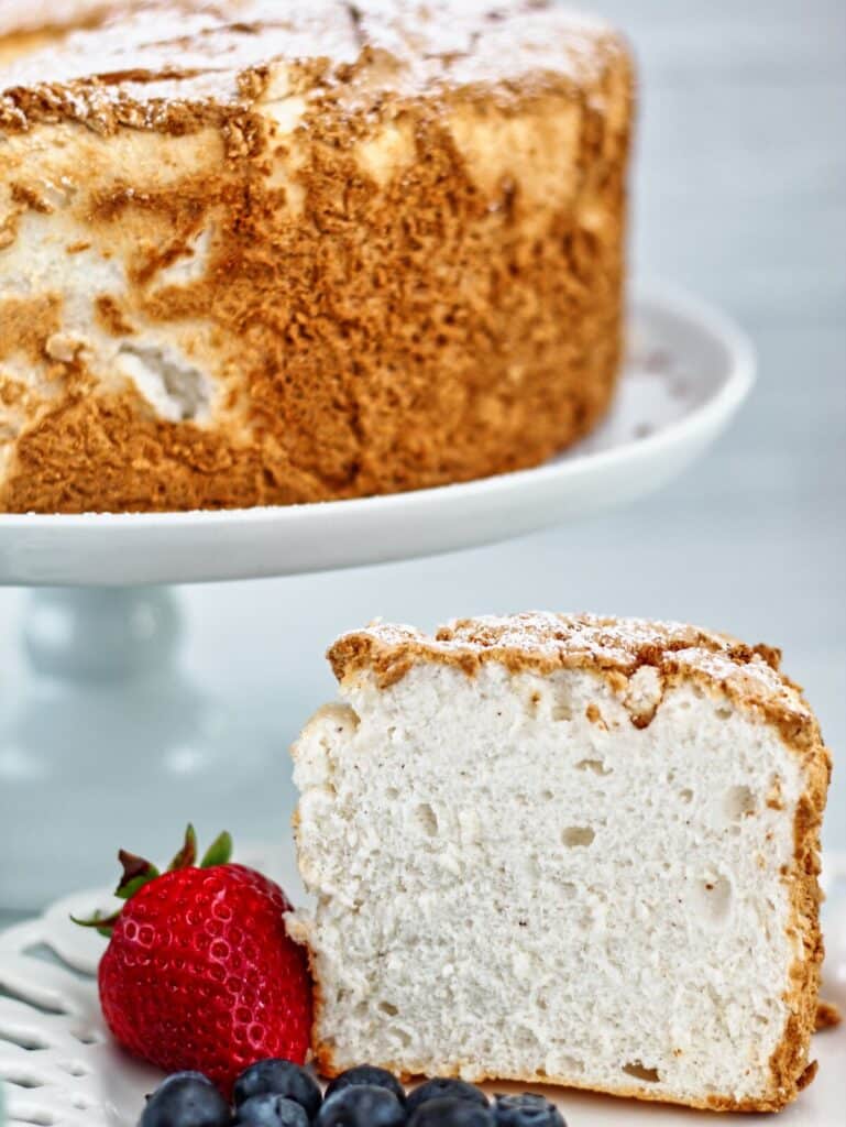 slice of angel food cake standing up straight on white plate with berries and whole cake in the background.