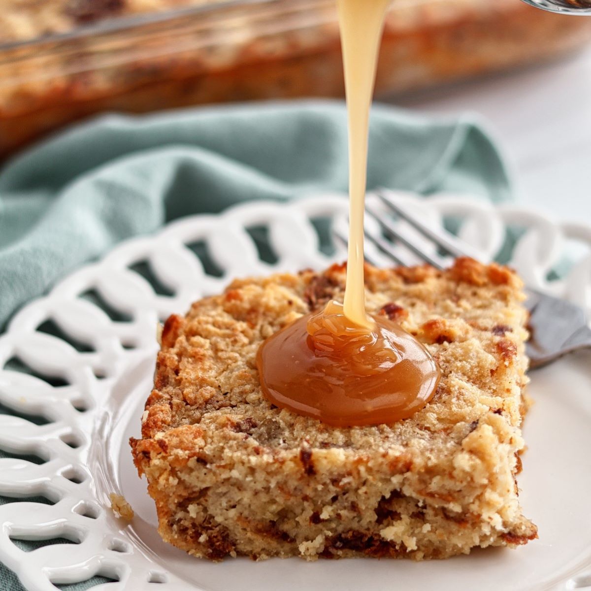 slice of gf bread pudding on white scalloped plate with sauce pouring over top.