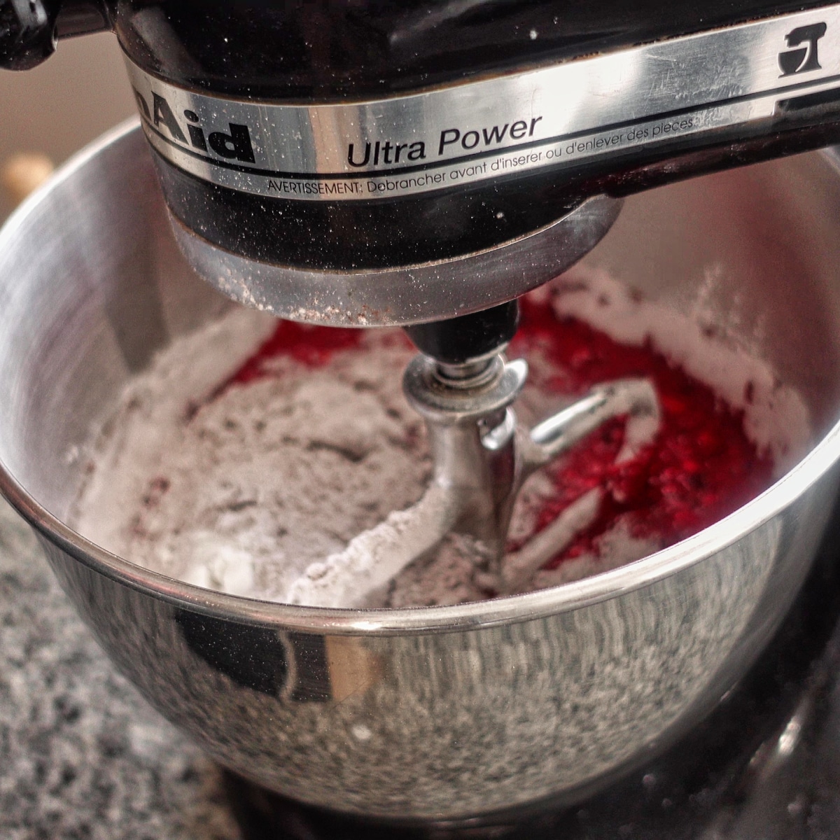 Adding dry ingredients and red food coloring to wet ingredients in stand mixer bowl.