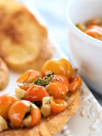 widescreen view of bruschetta on white platter with white bowl of marinated tomatoes alongside