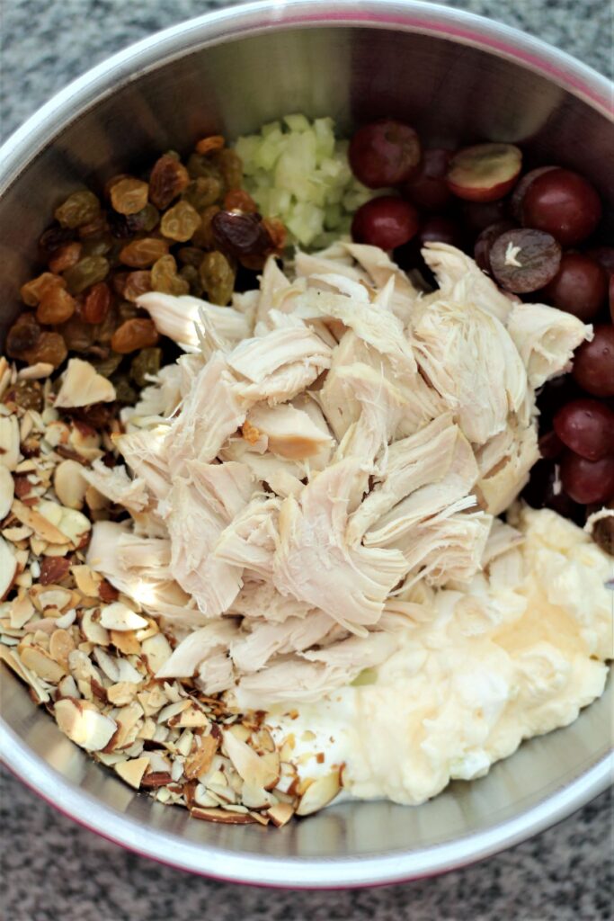 shredded chicken, sliced almonds, grapes, golden raisins, celery, mayonnaise, and sour cream in stainless steel bowl