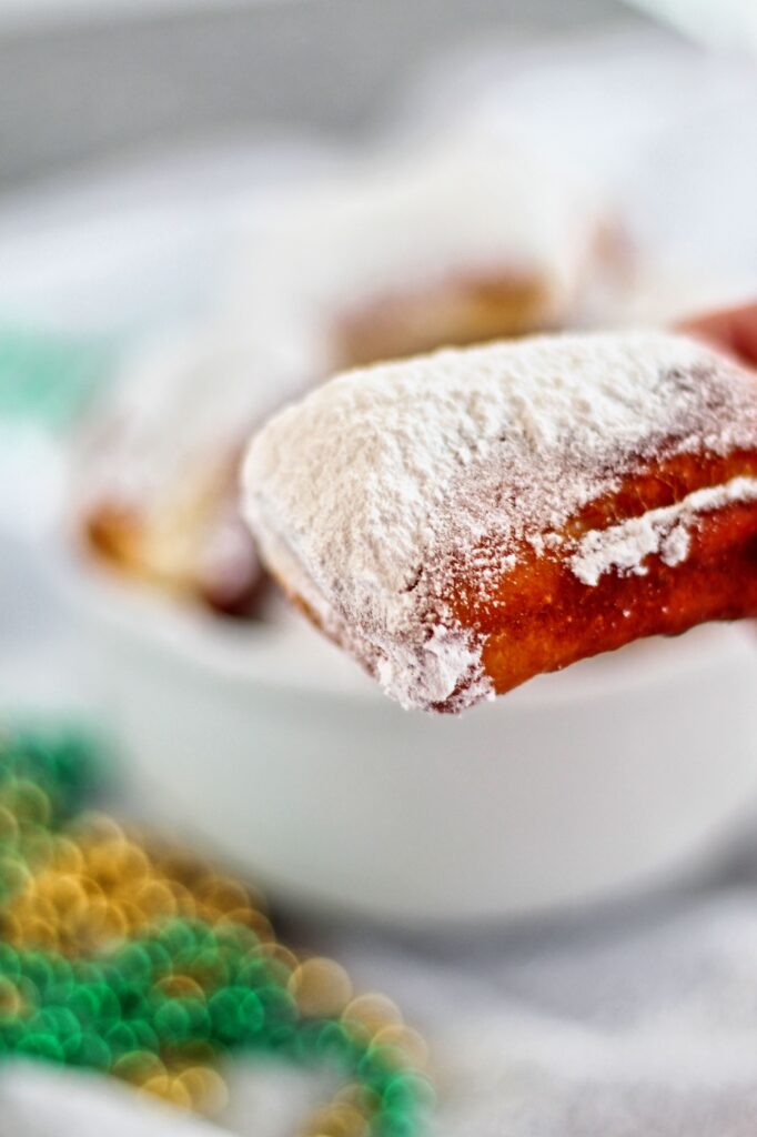 lots of powdered sugar piled onto a beignet with a bowl full of beignets in the background