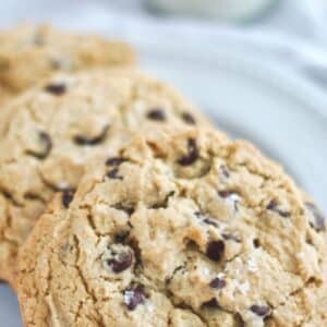 three large gluten free chocolate chip cookies on a white plate with milk in the background