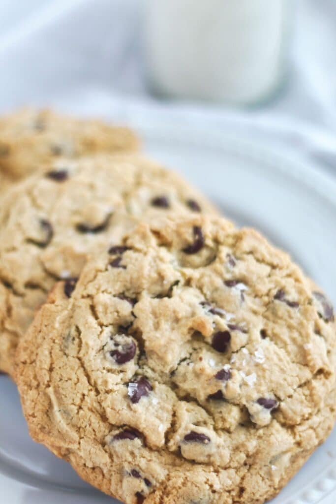 three large gluten free chocolate chip cookies on a white plate with milk in the background