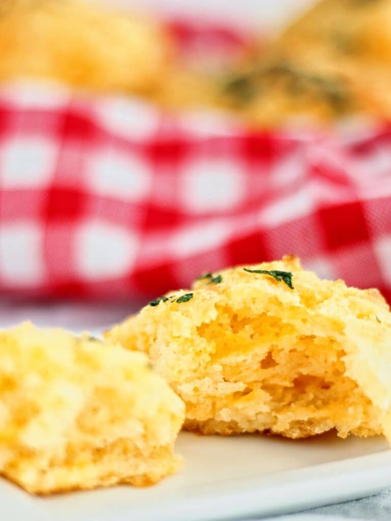 cheddar bay biscuit broken in half on small square plate in front of basket of biscuits in red checked napkin.