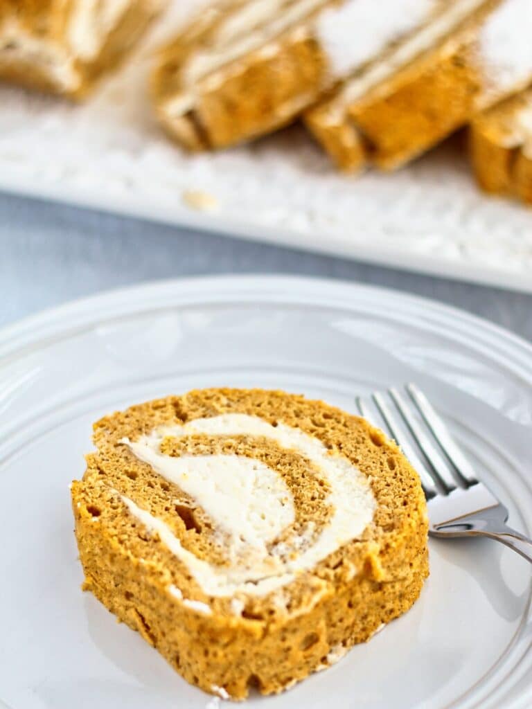 piece of pumpkin roll on white plate with platter of whole sliced pumpkin roll in background.