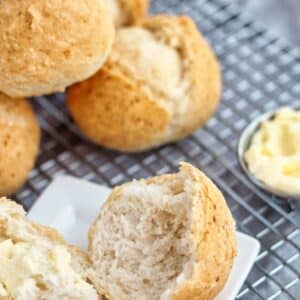 gluten free crusty french rolls on cooling rack with one roll split open on a small white square plate and butter to the side.