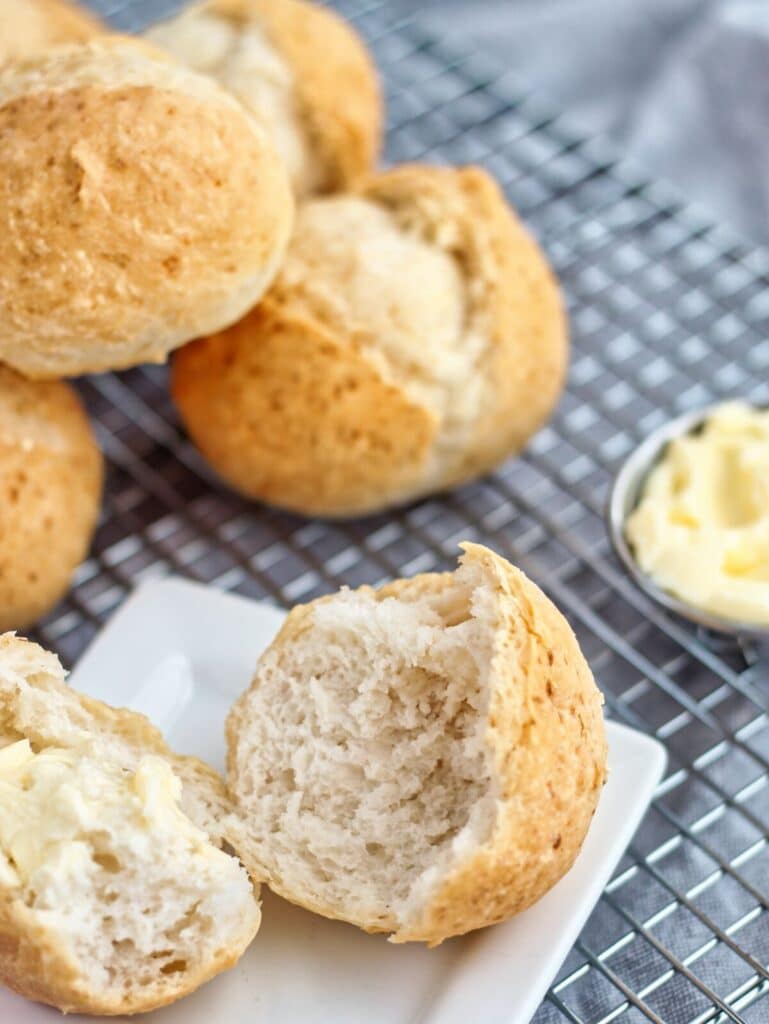 gluten free crusty french rolls on cooling rack with one roll split open on a small white square plate and butter to the side.