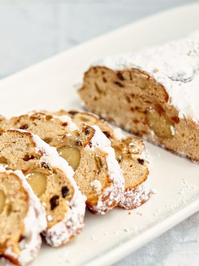 slight side angle view of a loaf of stollen on a rectangular white platter with half of it cut into half inch thick pieces.