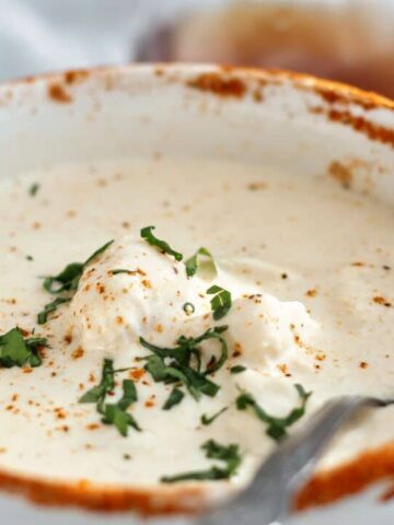 landscape view of Old Bay rimmed bowl of she crab soup on white linen with navy blue cloth napkin on the side and glass bottle of sherry in the background.