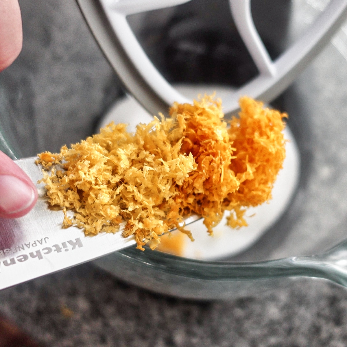 lemon zest on knife being scraped into glass mixer bowl with sugar in it.