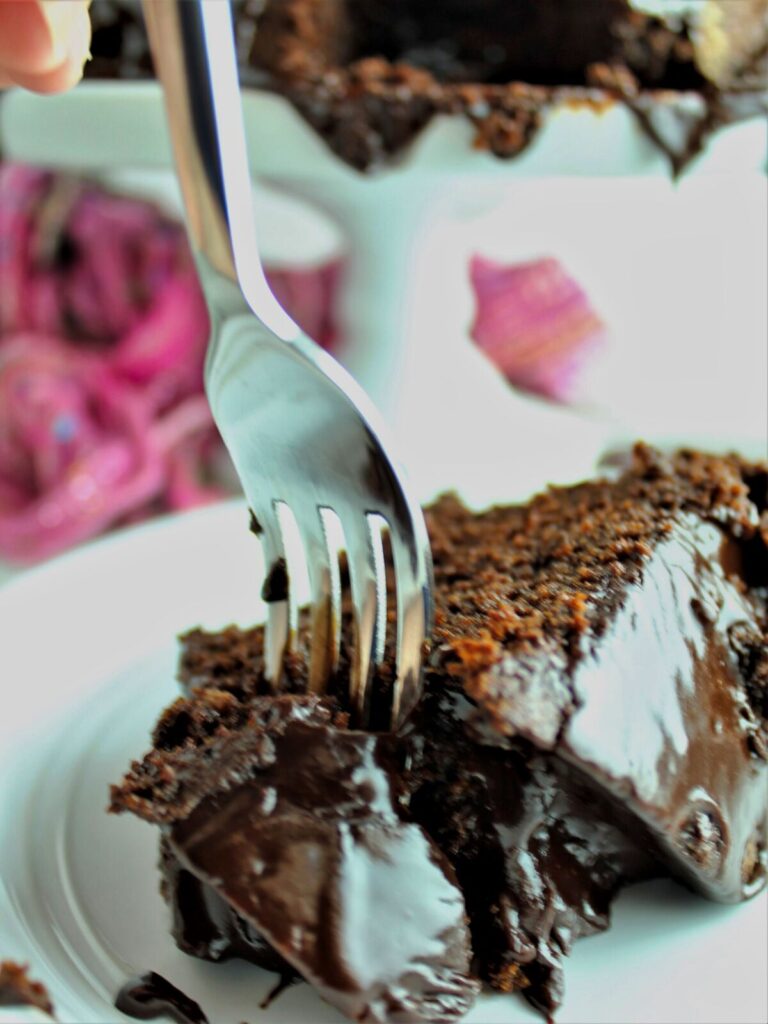 fork slicing through piece of chocolate fudge cake on white plate with rest of cake in the background on pedestal cake stand.