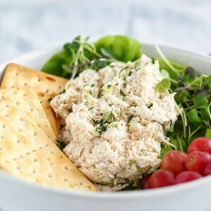 classic chicken salad on a bed of lettuce in a white bowl with grapes and crackers on the side.