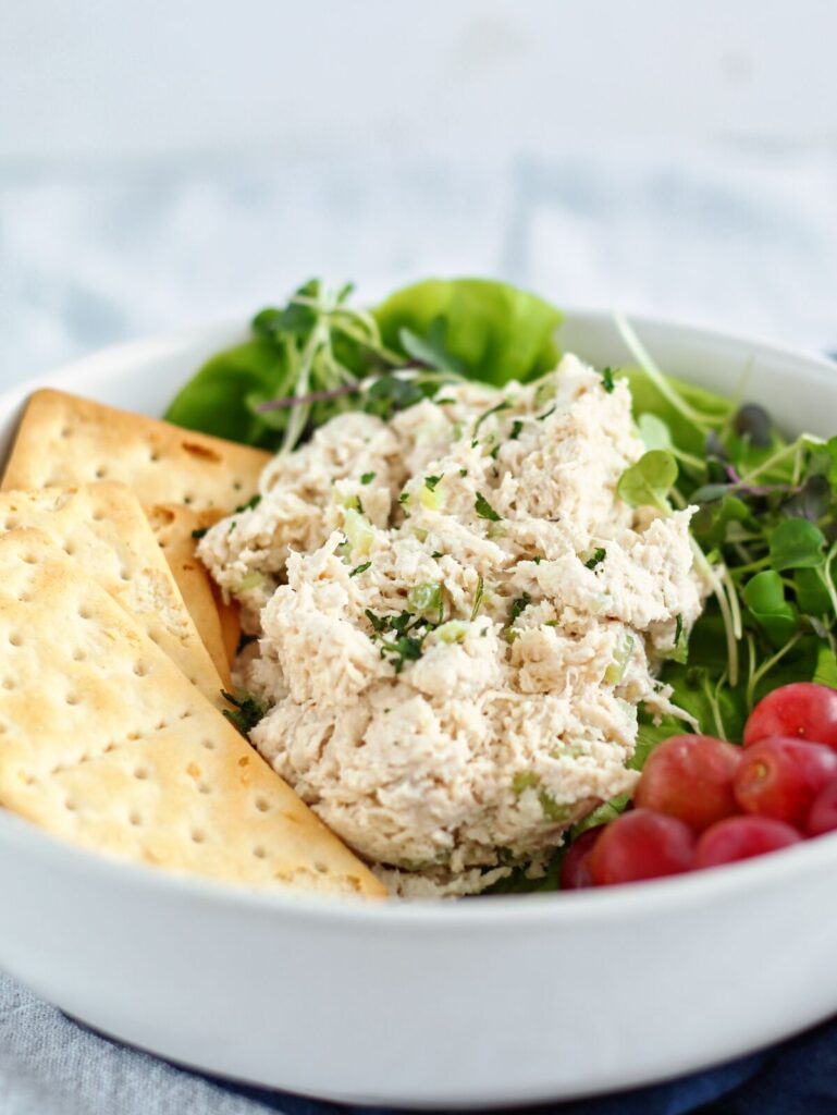 classic chicken salad on a bed of lettuce in a white bowl with grapes and crackers on the side.