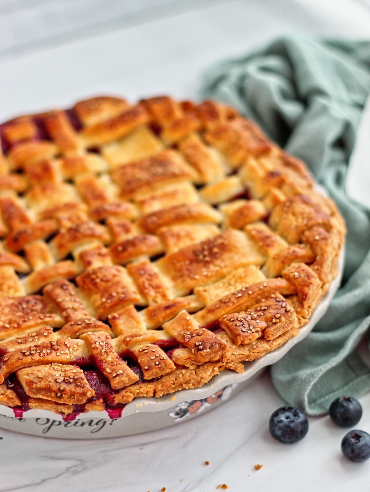 gluten free blueberry lattice top pie on white sandstone with light green towel to the side and stray blueberries.