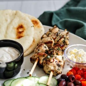 white platter with chicken skewers, pita, tahini sauce, cucumbers, olives, tomatoes, and feta.