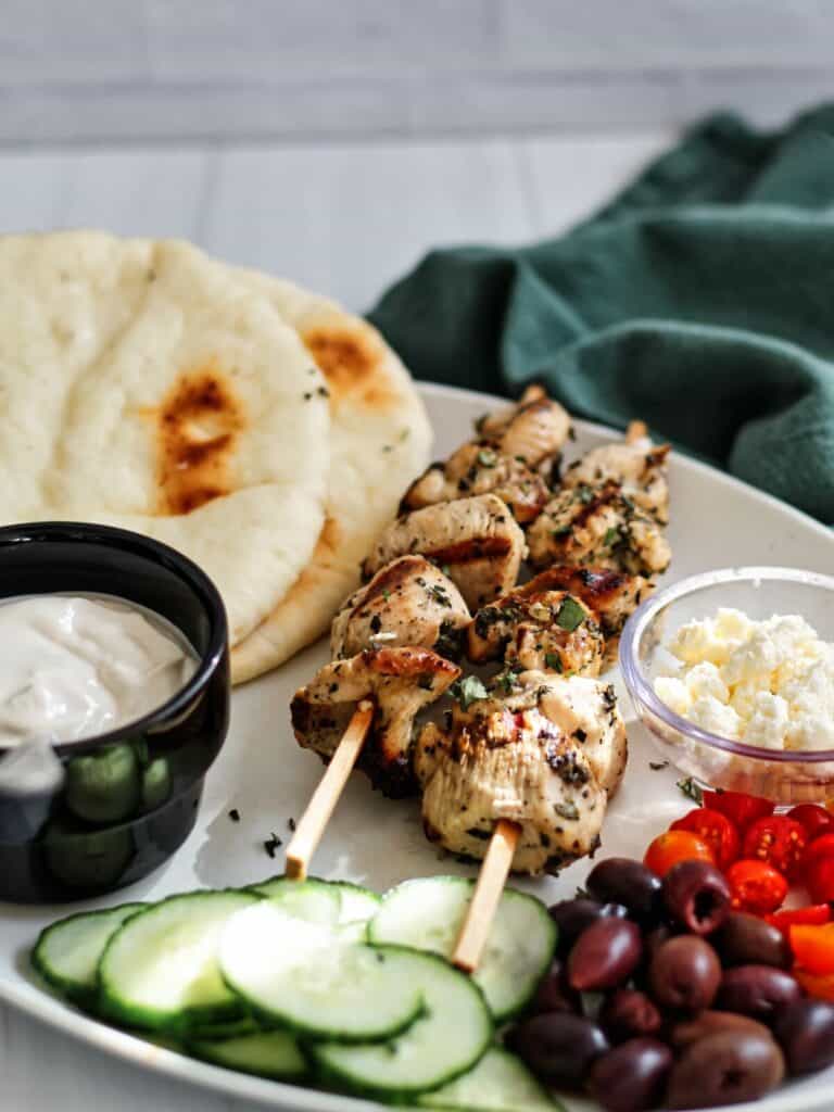 white platter with chicken skewers, pita, tahini sauce, cucumbers, olives, tomatoes, and feta.