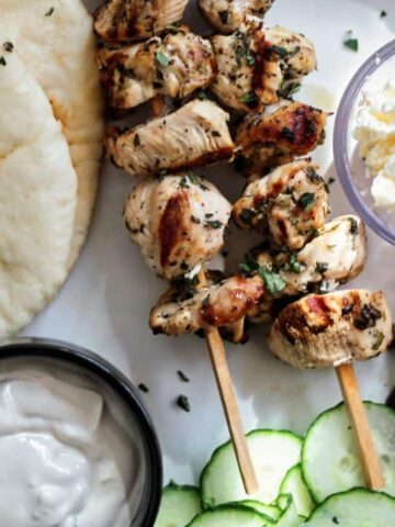 landscape view of grilled chicken souvlaki skewers on white platter with pita, tahini sauce, cucumber slices, kalamata olives, tomatoes, and feta cheese.