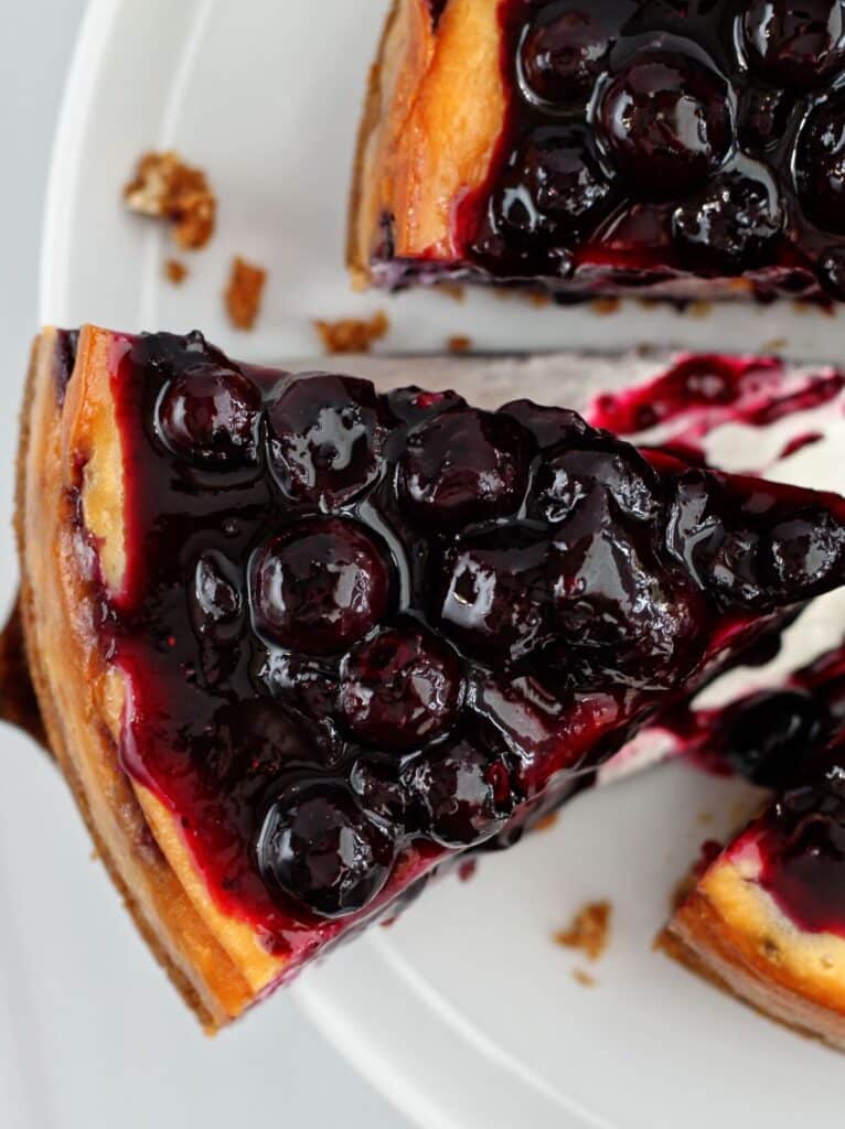 overhead view of one slice of blueberry cheesecake on serving spatula apart from whole cheesecake.