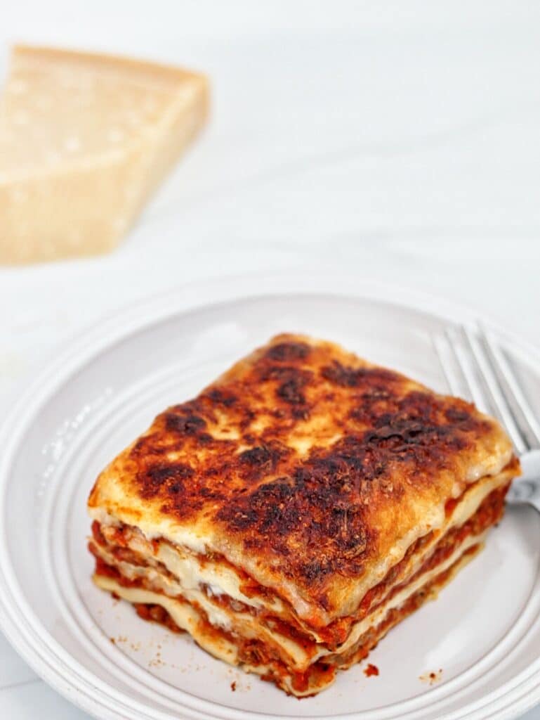 slice of lasagne on white plate with a hunk of parmesan cheese in the background.