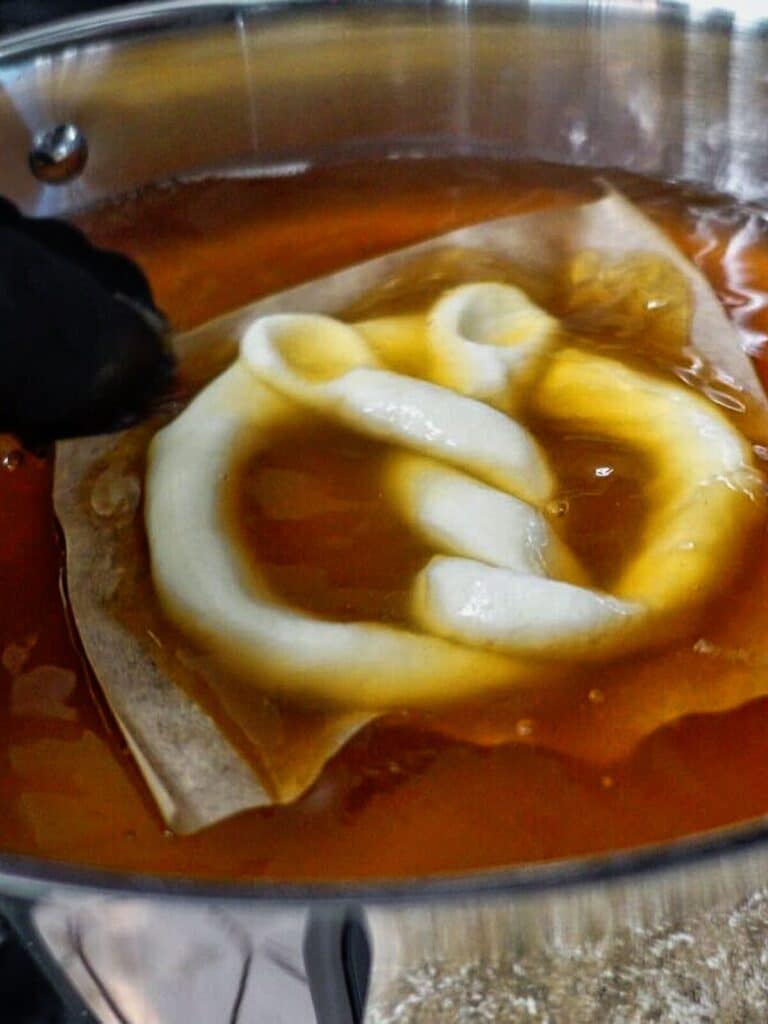 boiling pretzel in baking soda and molasses on parchment paper square.