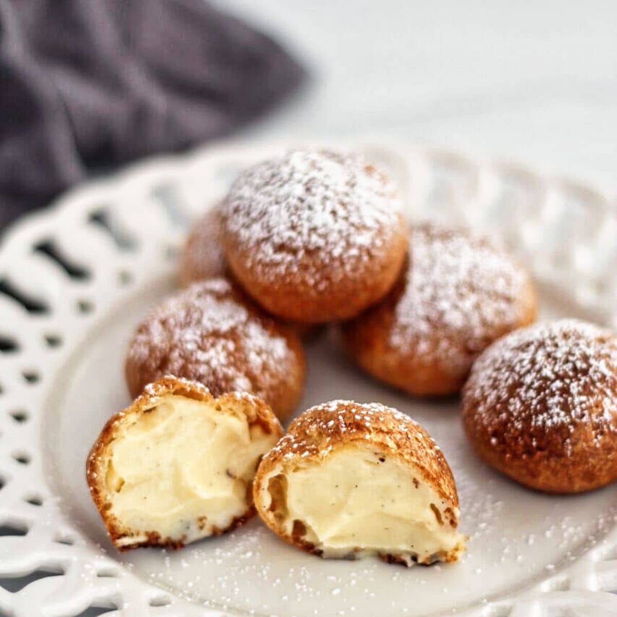 powdered sugar dusted cream puffs on white scalloped plate with cross section of one showing pastry cream.