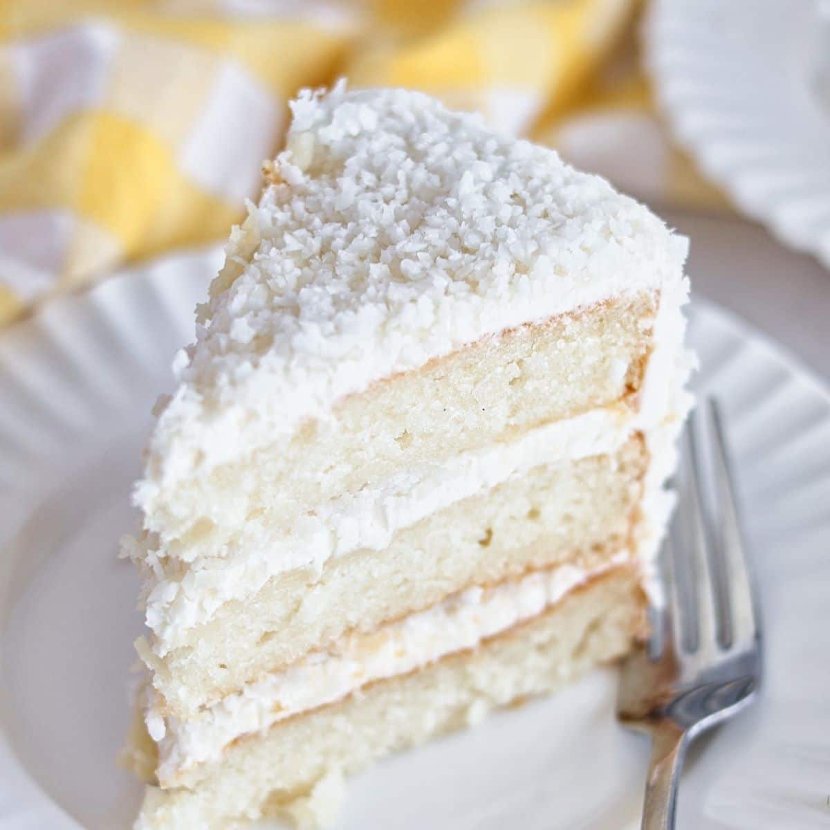 slice of coconut cake on white scalloped plate with yellow checked towel in the background.