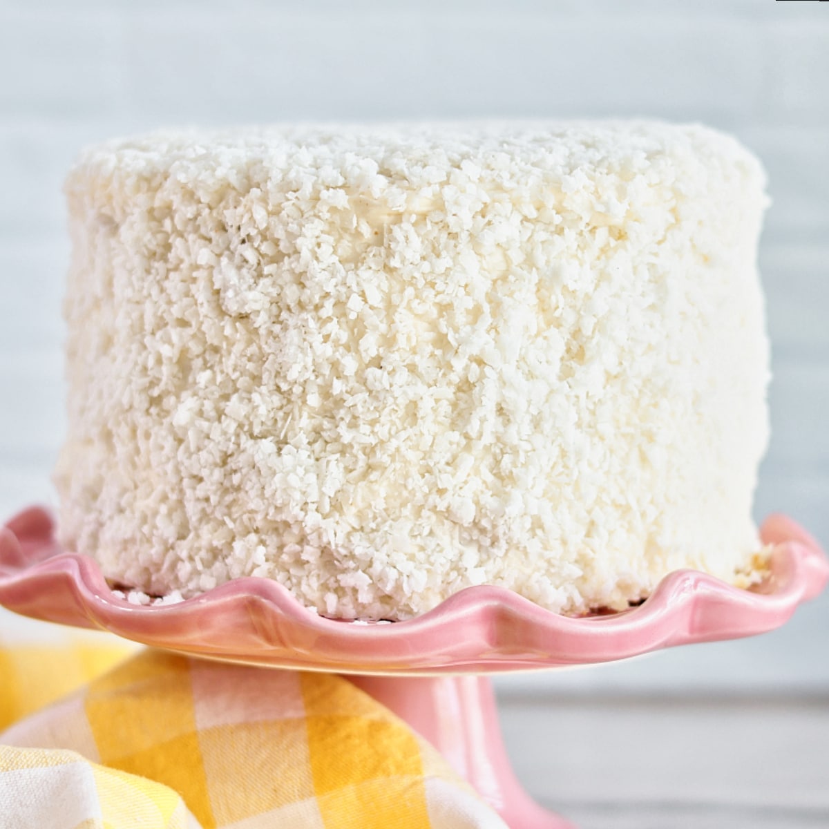 whole coconut cake on a pink cake stand with yellow checked towel on the side.