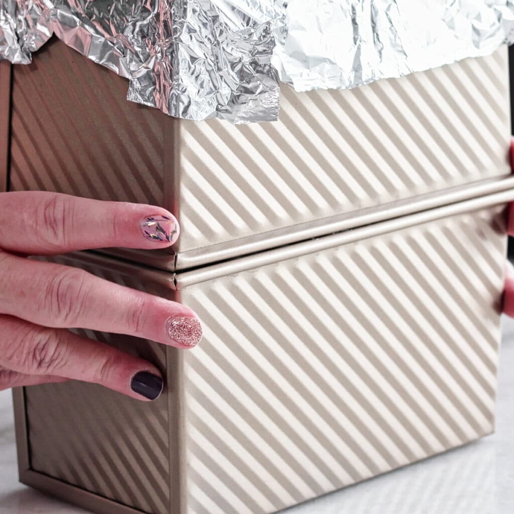 placing another loaf pan with foil on it on top of loaf pan with bread in it.