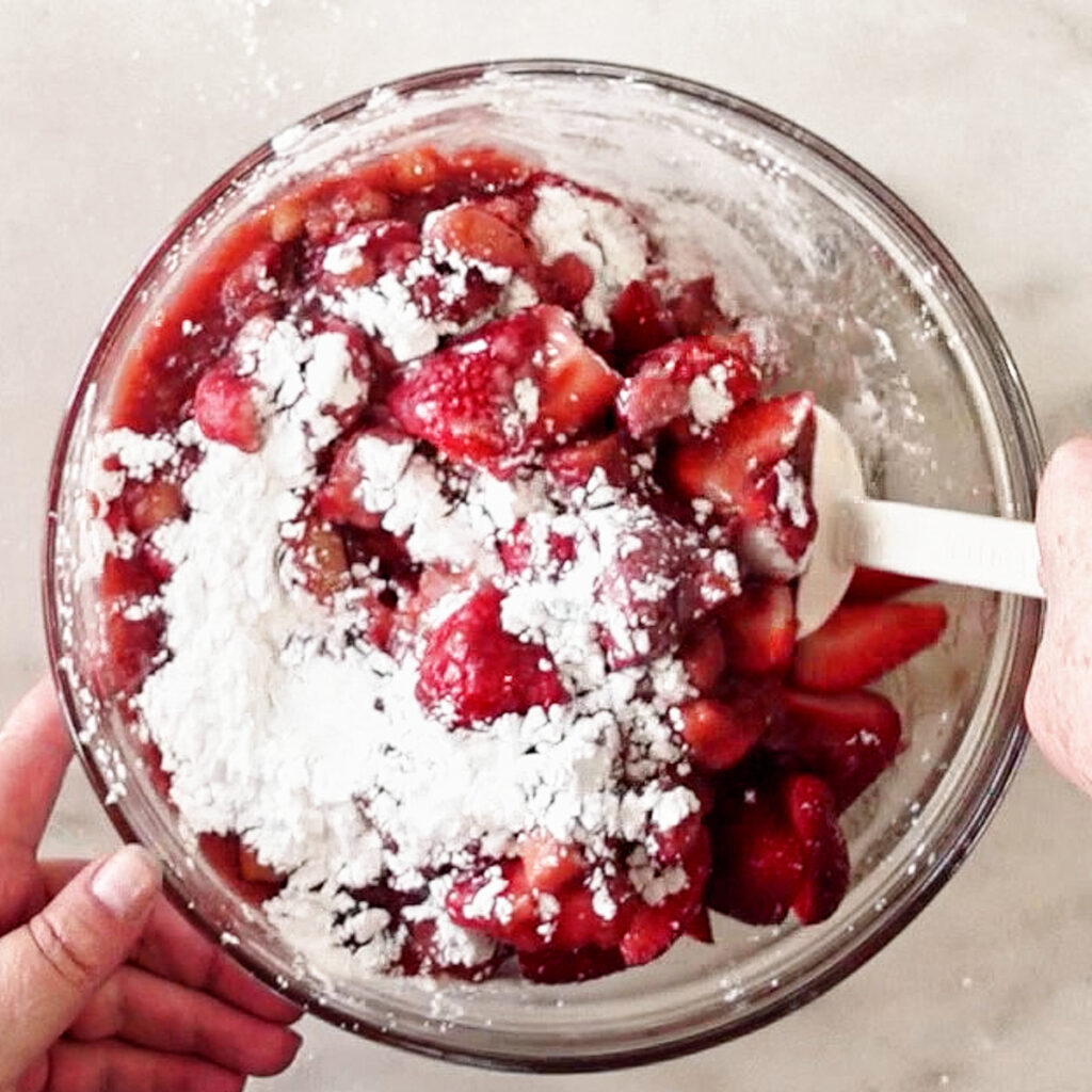 adding tapioca starch to strawberries and rhubarb in glass bowl and mixing with white spatula.