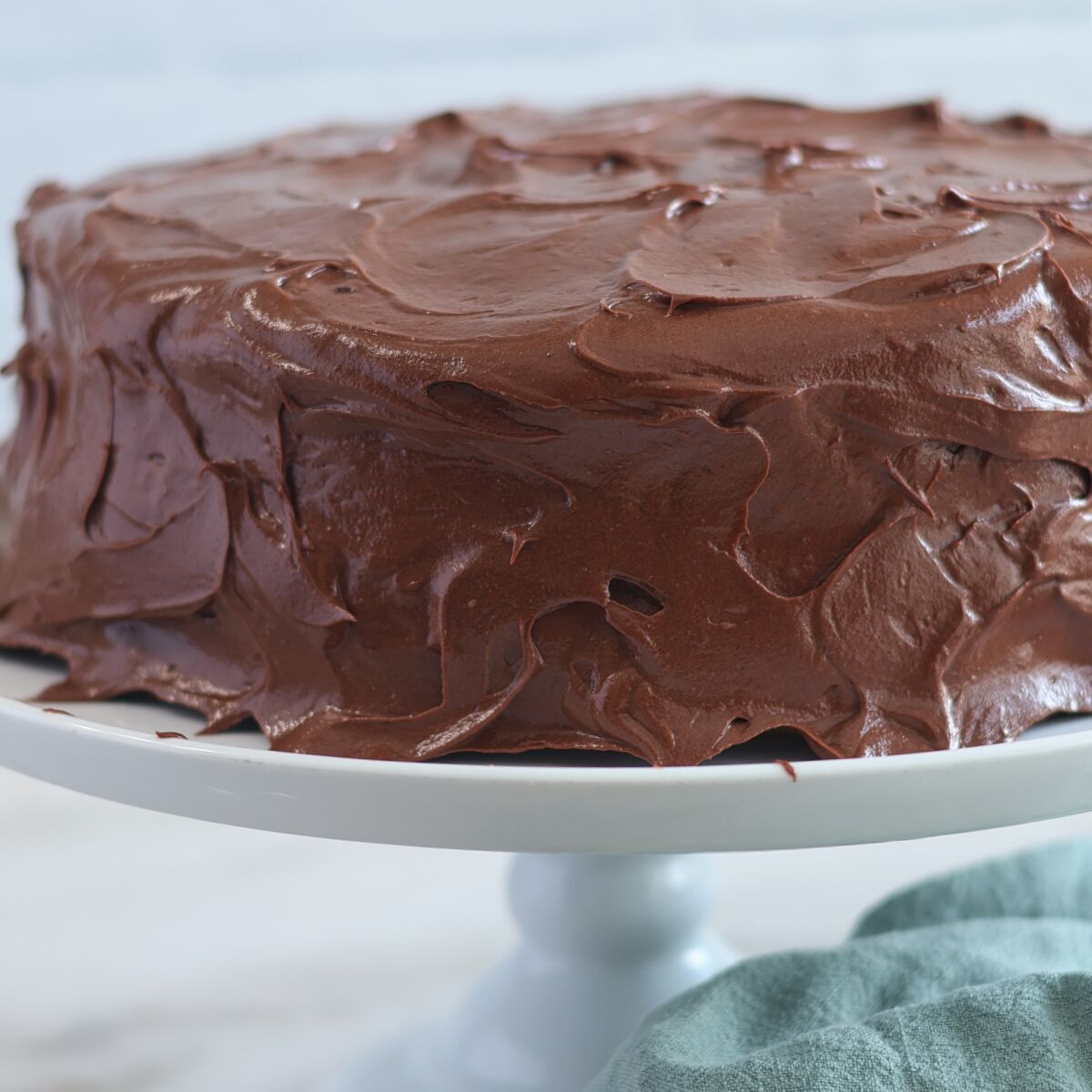 whole chocolate frosted cake on white cake stand with green towel on the counter.