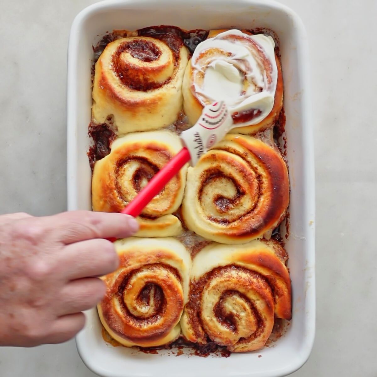 smearing cream cheese frosting onto one baked cinnamon roll.