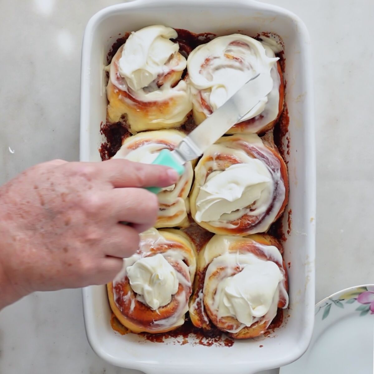 spreading final layer of icing onto rolls in white rectangular baking dish.
