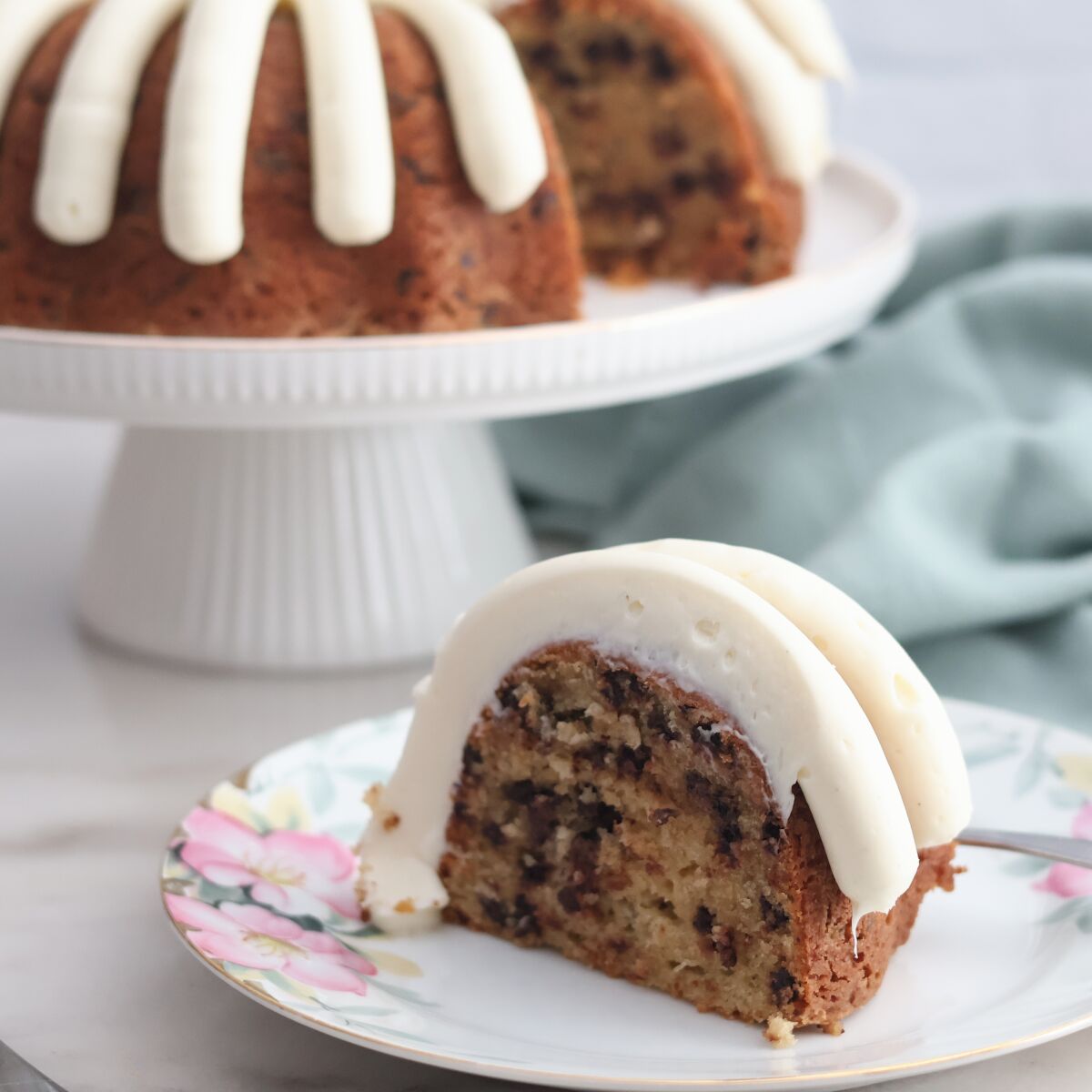 slice of cake on flowered plate with whole cake in the background on white cake stand.