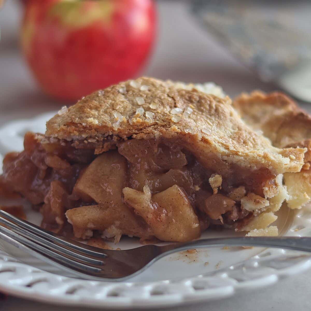 apple pie slice on white scalloped plate with apple in the background.