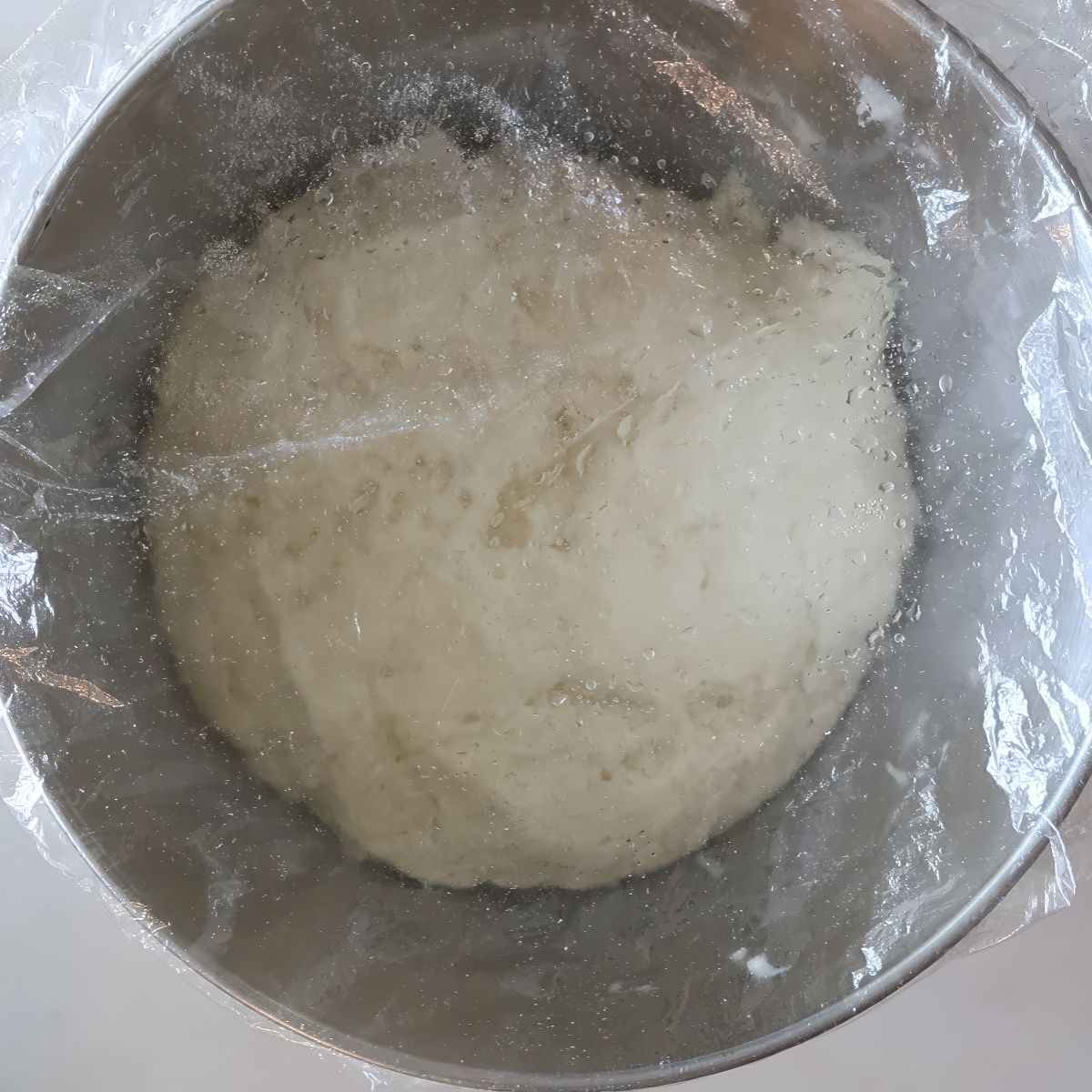 risen dough in stainless steel bowl covered with plastic wrap.